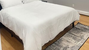Un dormitorio con cama, una mesita de noche, una ventana con persianas y una alfombra estampada.