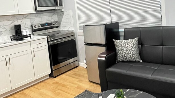 Cocina moderna con armarios blancos, microondas, estufa y refrigerador. Hay un sofá negro con un cojín estampado y una mesita con un control remoto y una planta.