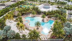 Vista aérea de un complejo turístico con piscina central, palmeras y cancha de tenis.