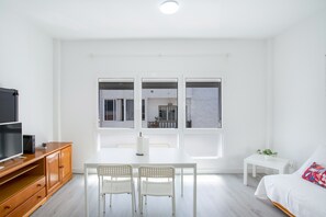 Ein modernes Zimmer mit Fernseher, Holzschrank, weißem Tisch mit Stühlen, einem kleinen weißen Beistelltisch und einem Bett mit farbigem Kissen.