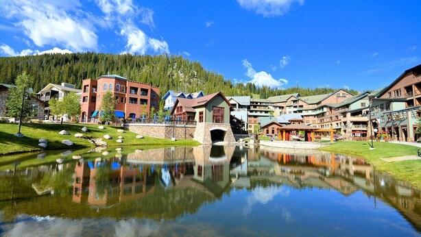 A lakeside resort with a central bridge, reflecting buildings, and a mountainous backdrop. - WP Resort Ski In Out Condo 2403 I Hot Tub I Gym I Ski Views I Bunk Room I Discounted Attractions (Winter Park)