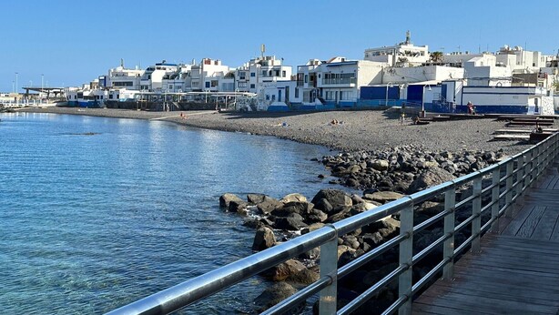 A coastal town with buildings along the waterfront, a rocky beach, and a wooden boardwalk with metal railings.