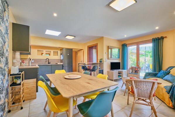 A modern kitchen with a dining area, a living room with a sofa and a TV, and a balcony with curtains.