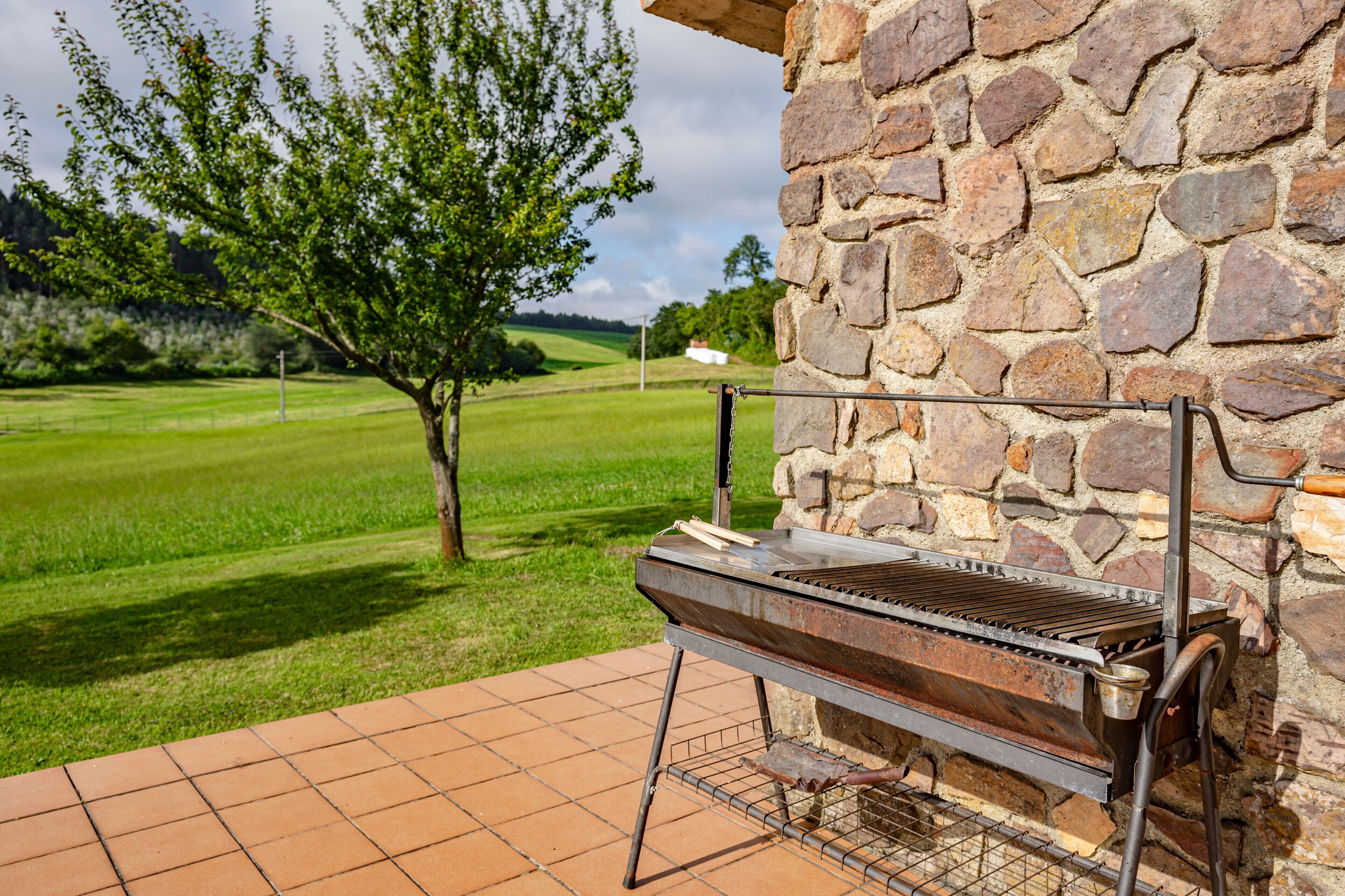 Un barbecue installé à l’extérieur, sur une terrasse, avec un mur de pierres en arrière-plan, un champ d’herbe et un arbre au loin.