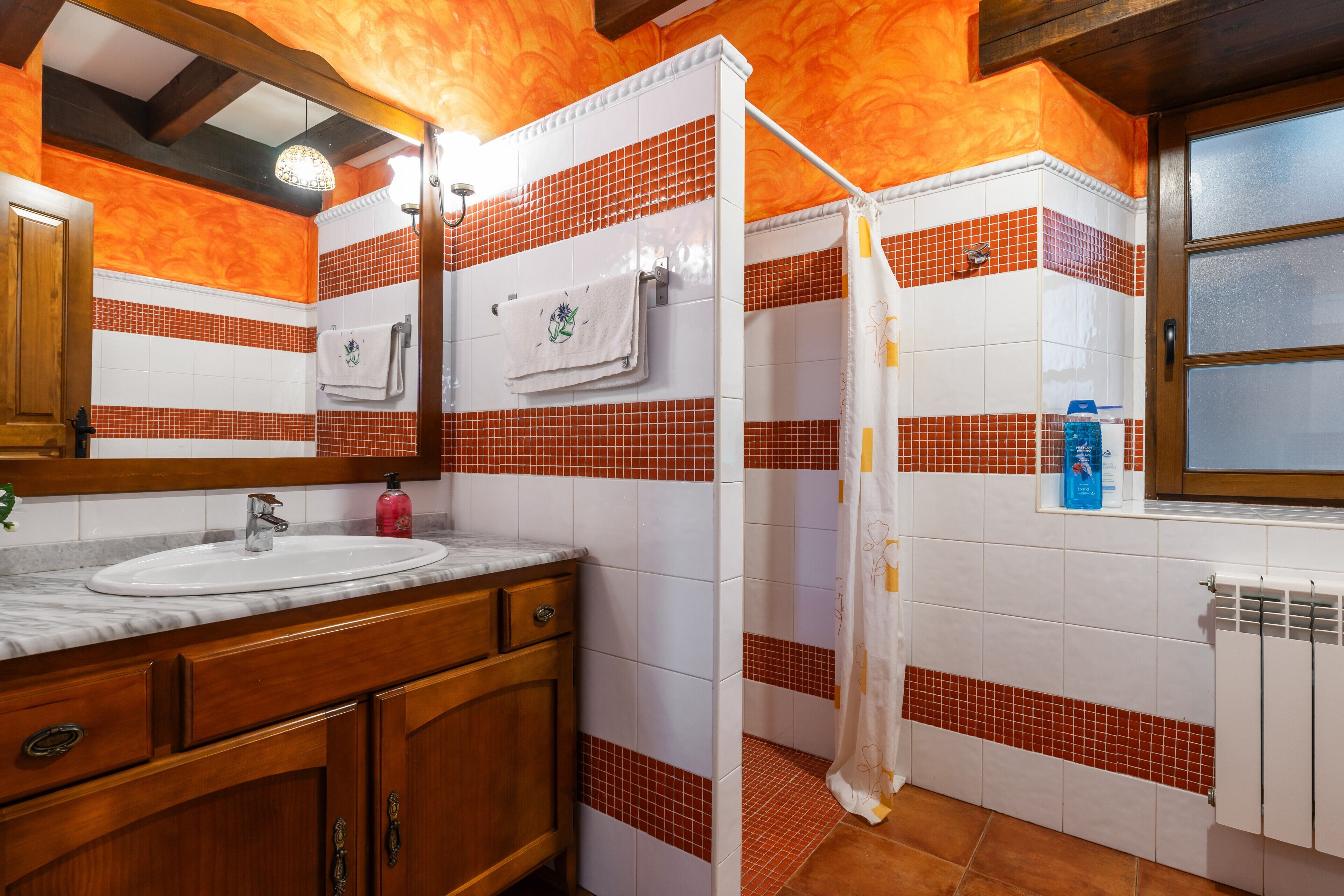 Ein Badezimmer mit einem weißen Waschbecken, einem Holz-Waschtisch, einem Spiegel und einer Duschkabine mit roten Fliesen.