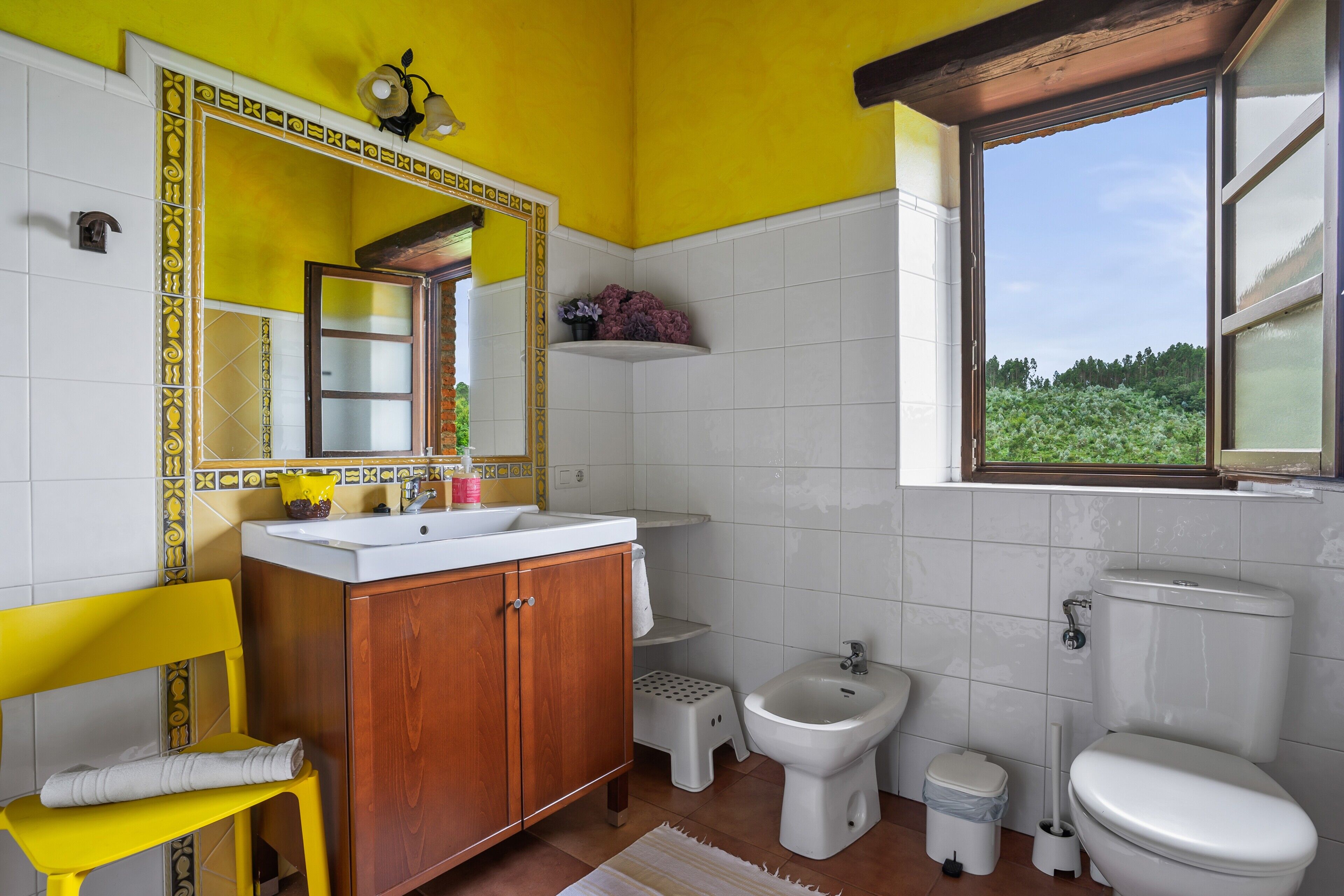 Un baño con pared amarilla, azulejos blancos, tocador de madera, una silla amarilla, un bidé y una ventana con vista a follaje.