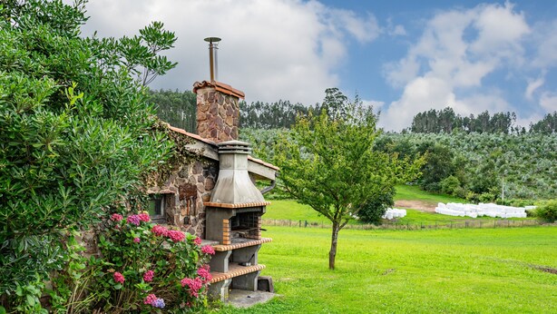 A rustic stone building with a chimney, surrounded by lush greenery and a clear sky. - Pedregal Farmhouse with Shared Terrace, Private Garden, and Wi-Fi (Gijon)