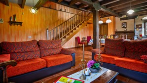 A cozy living room with a wooden staircase, two sofas with patterned cushions, a coffee table with a book and candle, and a dining area in the background. - Pedregal Farmhouse with Shared Terrace, Private Garden, and Wi-Fi (Gijon)
