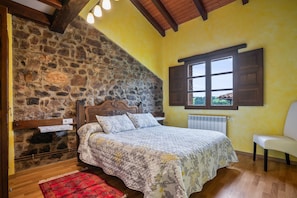 Quarto com parede de pedra, vigas de madeira no teto, cama com colcha florida, tapete vermelho e janela com persianas.