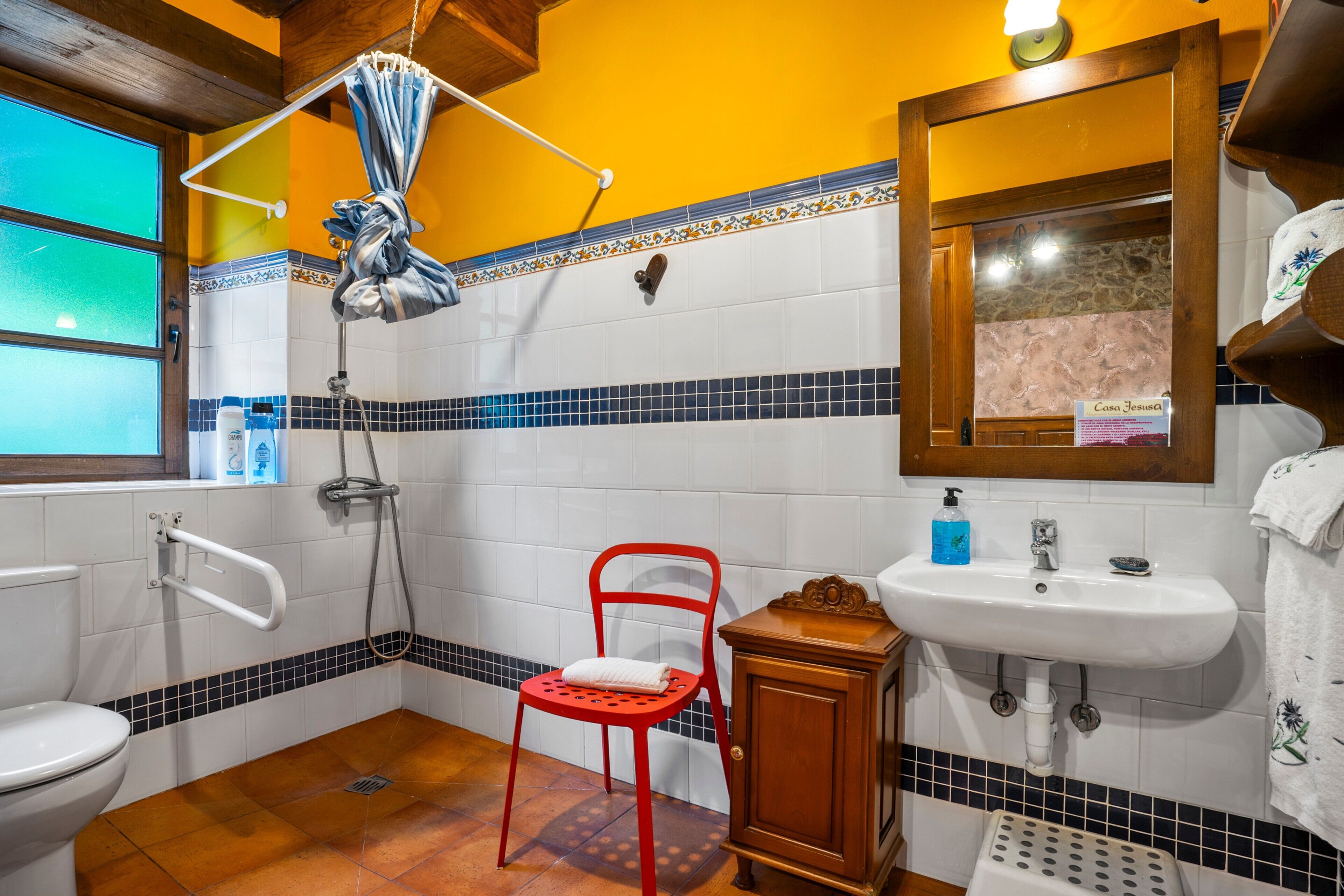 Une salle de bain avec des toilettes blanches, une chaise rouge, un meuble en bois, un lavabo et une douche avec un rideau.
