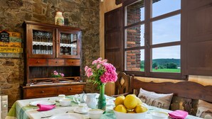 Une table à manger dressée avec des assiettes, des bols et un vase de fleurs, un buffet en bois avec des portes en verre, et une fenêtre donnant sur un paysage verdoyant.