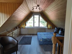 Un dormitorio acogedor en el ático, con techo inclinado, un ventanal grande, cama, silla y cómoda.