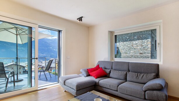 A modern living room with a grey sofa, red cushions, a glass coffee table, and a view of the sea through large glass doors.