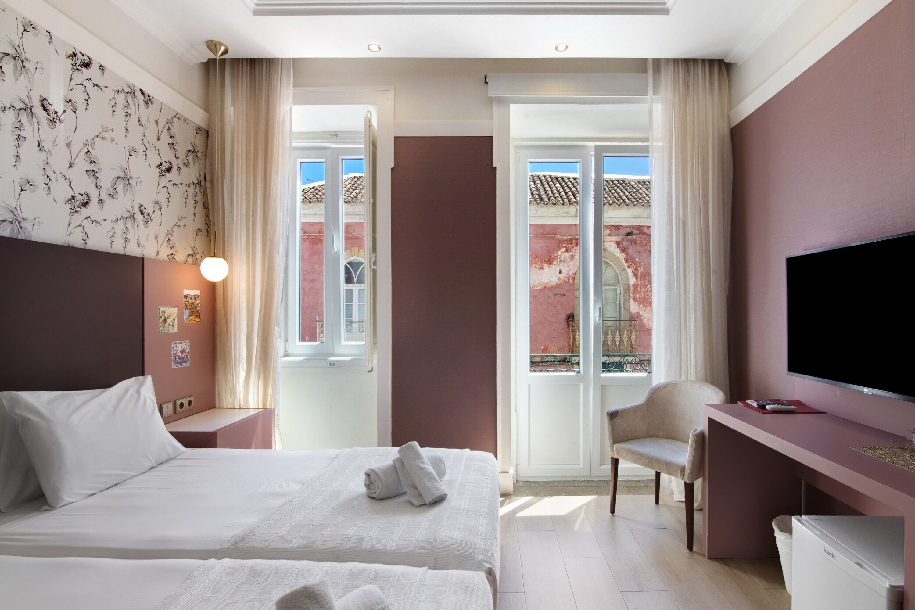 Une chambre d’hôtel avec deux lits, un bureau, une télévision et une vue sur un immeuble rouge, visible à travers une porte vitrée.