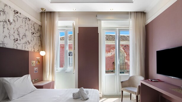 A hotel room with two beds, a desk, a TV, and a view of a red building through a glass door. - Room 'Quarto Twin Comfort Plus 101' with Shared Terrace, Wi-Fi and Air Conditioning (Faro)