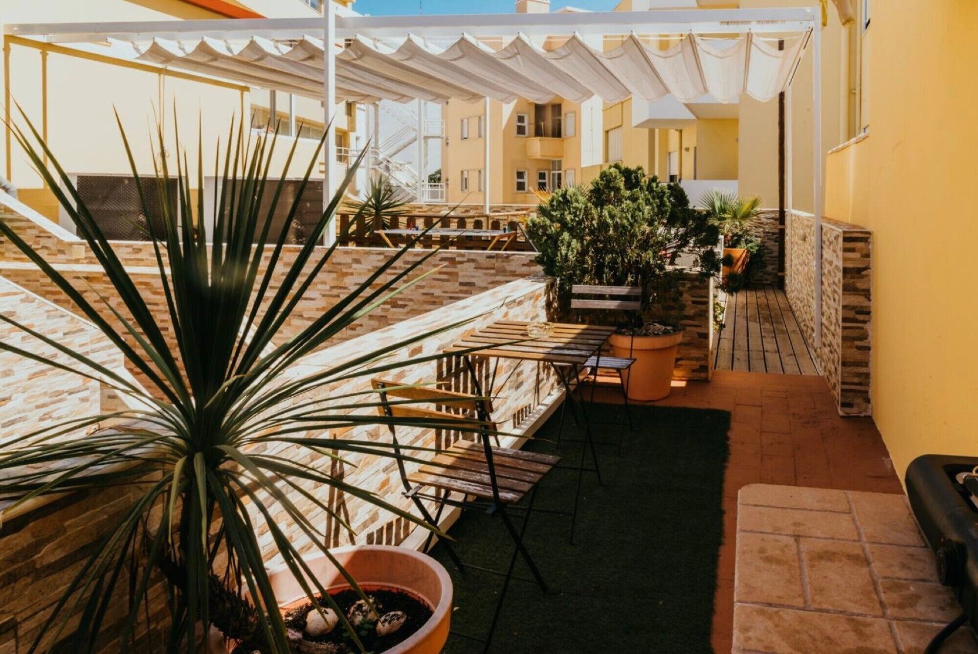 Une terrasse extérieure couverte, aménagée avec des meubles en bois, des plantes en pot et un mur en pierre.
