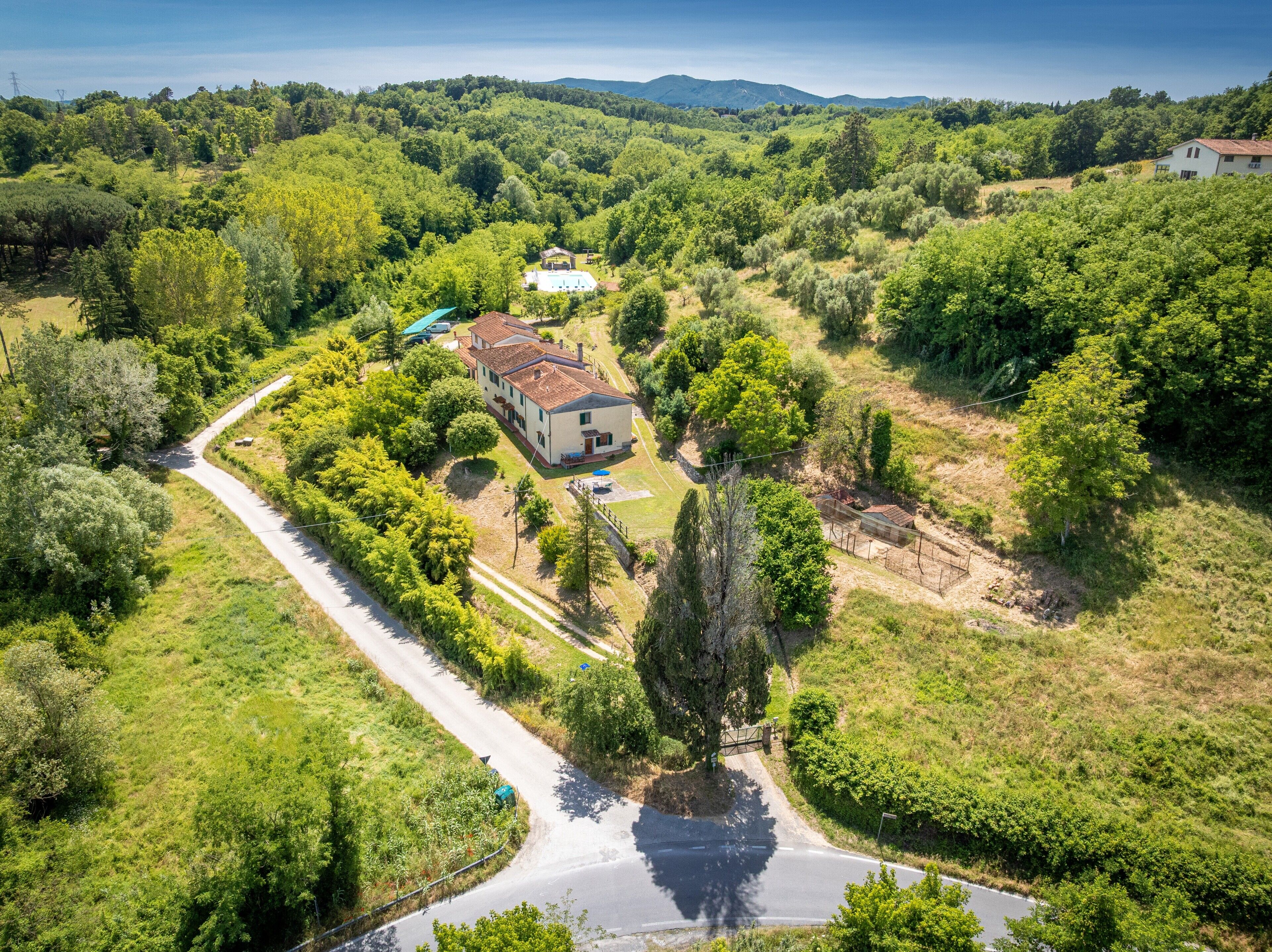 A rural house with a paved driveway, surrounded by dense greenery and hills in the background.