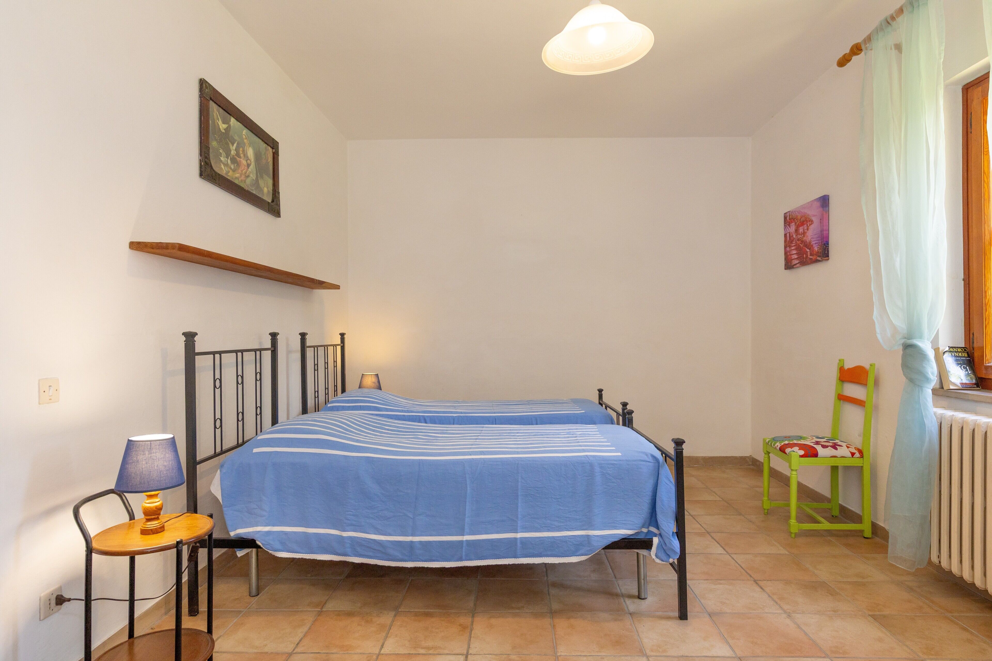 A bedroom with a bed, a chair, a table with a lamp, a shelf, and a radiator.