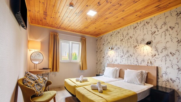 Ein Hotelzimmer mit Holzboden, zwei Betten mit gelber Bettwäsche, einem Sessel, einem Nachttisch, einer Lampe, einem Fenster mit Vorhängen und floralem Wandpapier.