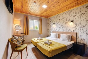 Habitación de hotel con techo de madera, dos camas con ropa de cama amarilla, una silla, una mesita de noche, una lámpara, una ventana con cortinas y papel tapiz con motivos florales.