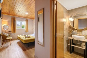 Une pièce avec un plafond en bois, un lit avec une literie jaune, une chaise, un bureau, une fenêtre avec des rideaux et une salle de bain avec un lavabo et un miroir.