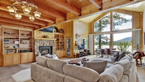 Un salón espacioso con un amplio sofá modular, estanterías de madera, una chimenea de piedra y grandes ventanales que ofrecen vistas al exterior.