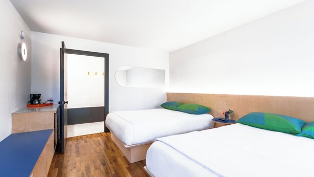 Ein Hotelzimmer mit zwei Betten, Holzboden, Schreibtisch mit Kaffeemaschine und einer Wandablage.