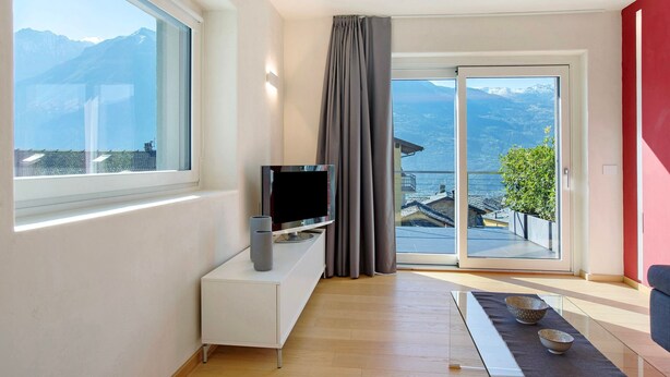 Una sala de estar moderna con un televisor sobre una consola blanca, una mesa de vidrio con cuencos y un ventanal corredero a un balcón con vista a la montaña.