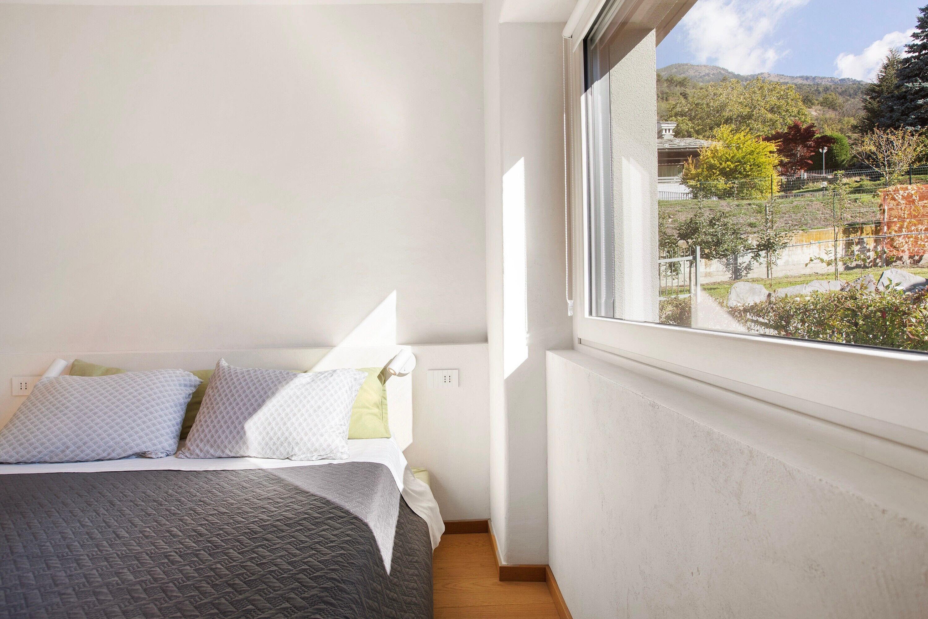 A bedroom with a bed, pillows, a window, and a view of a garden and buildings.
