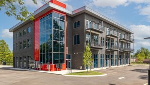 Een modern gebouw met een gevel in rood en grijs, grote ramen en een parkeerplaats.