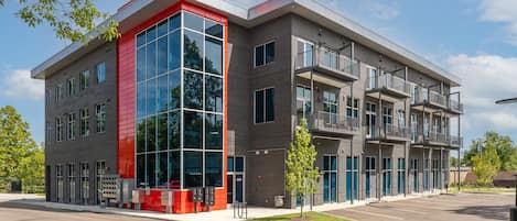A modern building with a red and grey facade, large windows, and a parking lot.