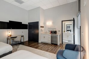 A modern hotel room with a bed, a desk with a chair, a kitchenette, and a bathroom visible through an open door. - Professional Retreat Double Queen Room (Nashville)