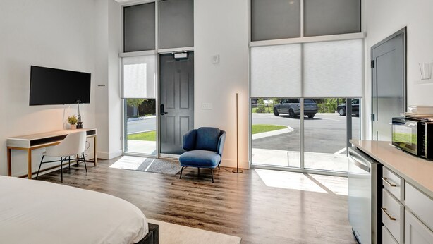 Ein modernes Hotelzimmer mit einem großen Bett, einem Schreibtisch mit Stuhl, einem Fernseher und Blick auf die Straße durch große Fenster.