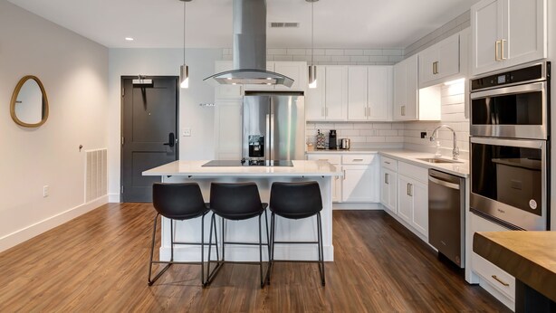 Una cocina moderna con armarios blancos, electrodomésticos de acero inoxidable, una isla central con taburetes y luces colgantes.