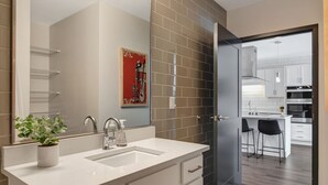 Un baño moderno con un lavamanos blanco, un espejo y una planta en maceta. Se ve una puerta que da a una cocina con armarios blancos y un taburete de bar.