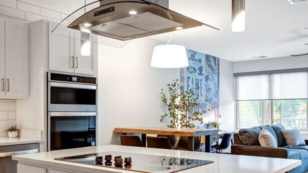 Una cocina moderna con armarios blancos, una isla central, electrodomésticos de acero inoxidable, iluminación colgante y vistas a una sala de estar con un sofá y una planta.