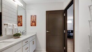 Un baño moderno con azulejos blancos, un espejo grande, dos obras de arte con temática de guitarra, una puerta marrón oscuro y un lavamanos blanco con detalles dorados.