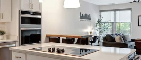 Une cuisine moderne avec des meubles blancs, un îlot central, des appareils en acier inoxydable, des luminaires suspendus et une décoration murale représentant un paysage urbain.