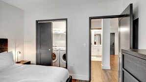 A modern hotel room with a bed, a nightstand, a lamp, a wardrobe, and a view into the bathroom with a washing machine. - Neon Nest: One Bedroom Nashville Getaway (Nashville)