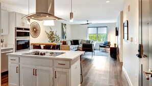 Een moderne keuken met witte kasten, een centraal eiland en roestvrijstalen apparatuur. Er is een eethoek met een tafel en stoelen, en een zithoek met een bank en een groot raam.