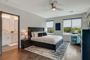 Un dormitorio moderno con una cama grande, un cabecero oscuro, una mesita de noche con una lámpara y vistas al exterior a través de una ventana.