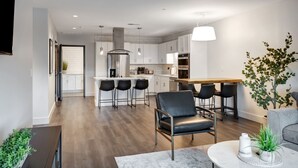 A modern kitchen with white cabinets, a central island, and bar stools. There is a dining area with a grey sofa and a coffee table.