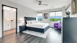 A modern bedroom with a large bed, a ceiling fan, and a view of the outdoors through the windows.