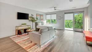 Un salon moderne avec un canapé, un téléviseur à écran plat, une petite table sur laquelle est posée une plante, un lit et une grande fenêtre donnant sur un paysage verdoyant.
