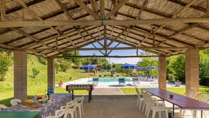 Un pavillon extérieur doté d’un toit de chaume, avec des tables, des chaises et une piscine en arrière-plan.