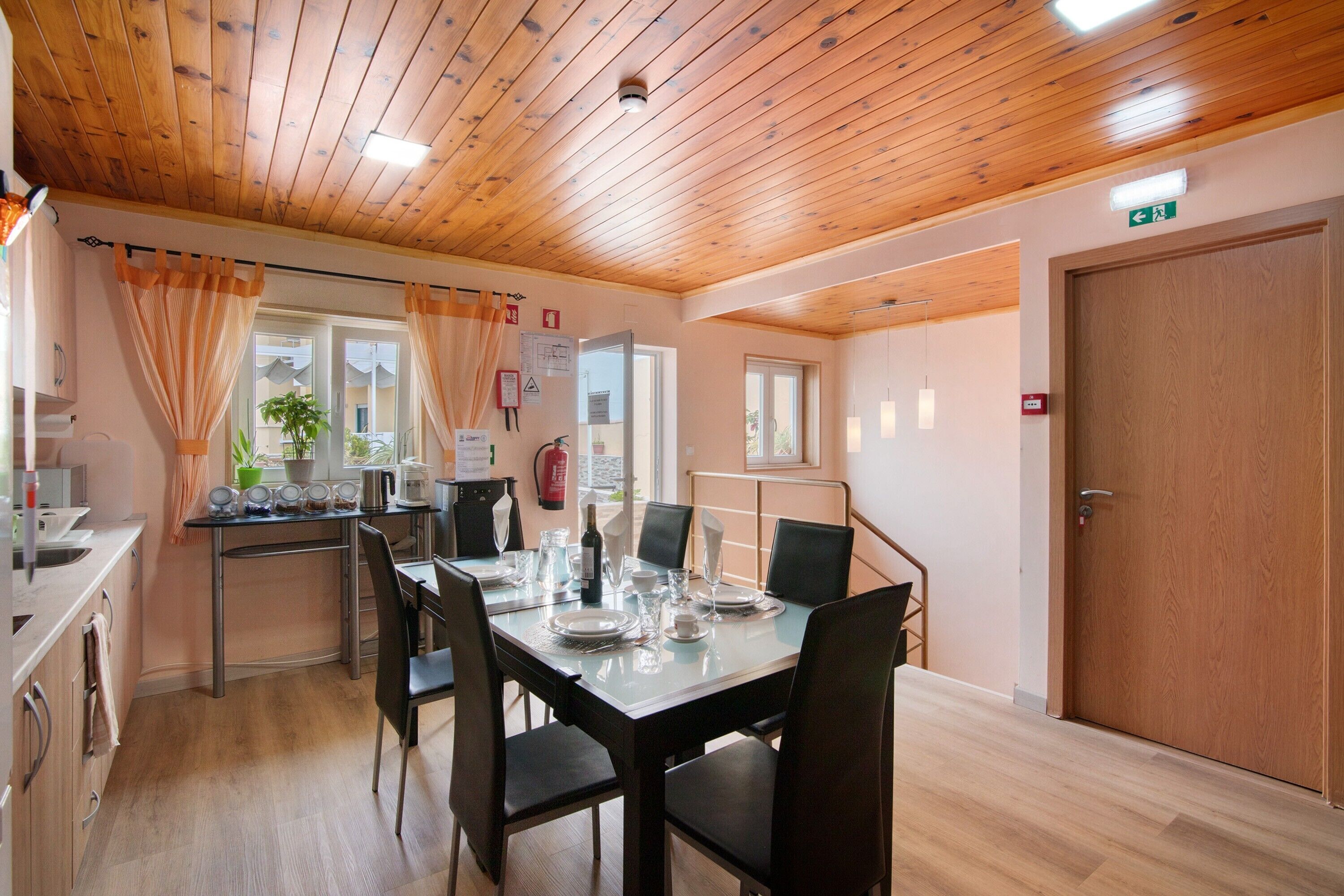 Un coin repas avec un plafond en bois, une table dressée pour quatre personnes et une porte portant un panneau indiquant la sortie de secours.