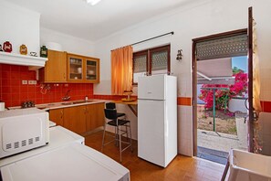 Cocina pequeña y funcional con refrigerador, microondas y lavamanos. Hay una ventana con cortinas, una silla y una mesita.
