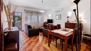 A living room with a sofa, a dining table with chairs, a TV, and a lamp. - Holiday Home 'Casa Maria Beatriz' with Private Terrace, Private Garden and Wi-Fi (Vila Nova de Cacela)