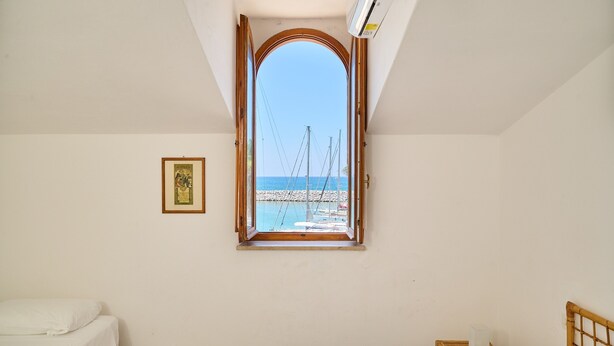A room with a bed, a small bedside table, a framed picture on the wall, and a window with a view of a pier and boats. - Holiday Home 'Villa Liberty Rosa' with Sea View, Wi-Fi and Air Conditioning (San Vincenzo)