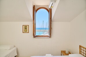 Een kamer met een bed, een klein nachtkastje, een ingelijst foto aan de muur, en een raam met uitzicht op een pier en boten.